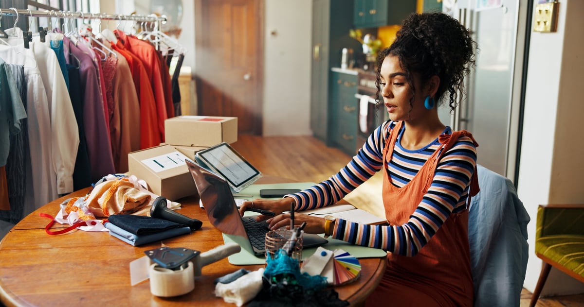 Fashion designer working on laptop in small business studio surrounded by fabric