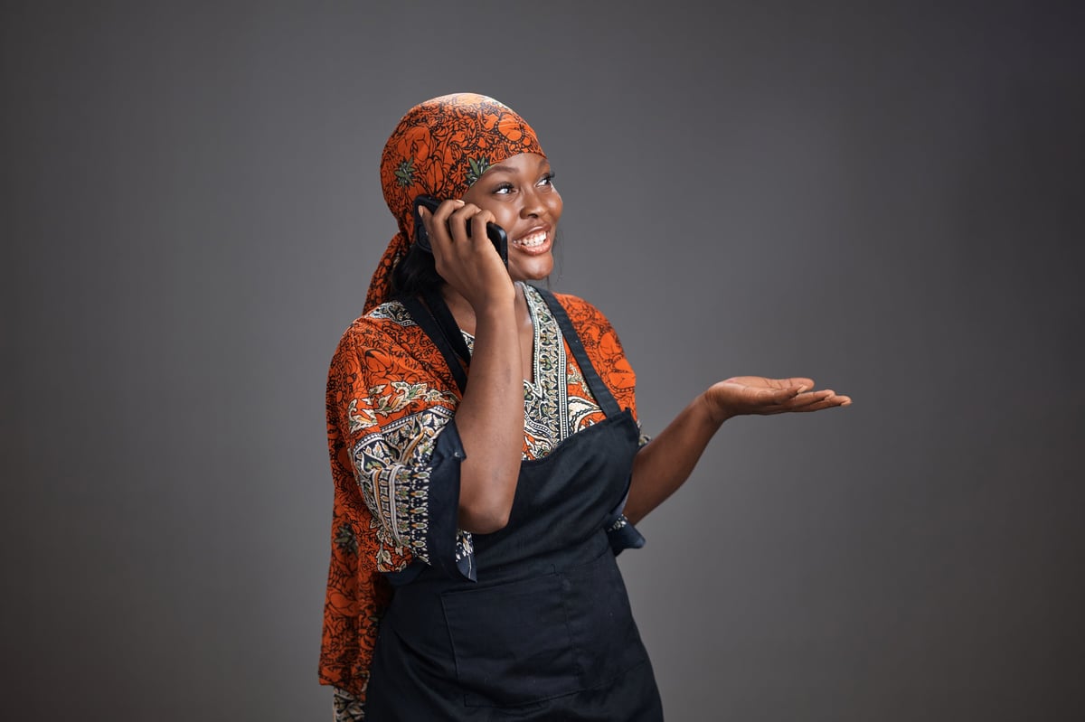 Young black woman wearing a patterned traditional dress, headscarf, and apron smiles while talking on a smartphone